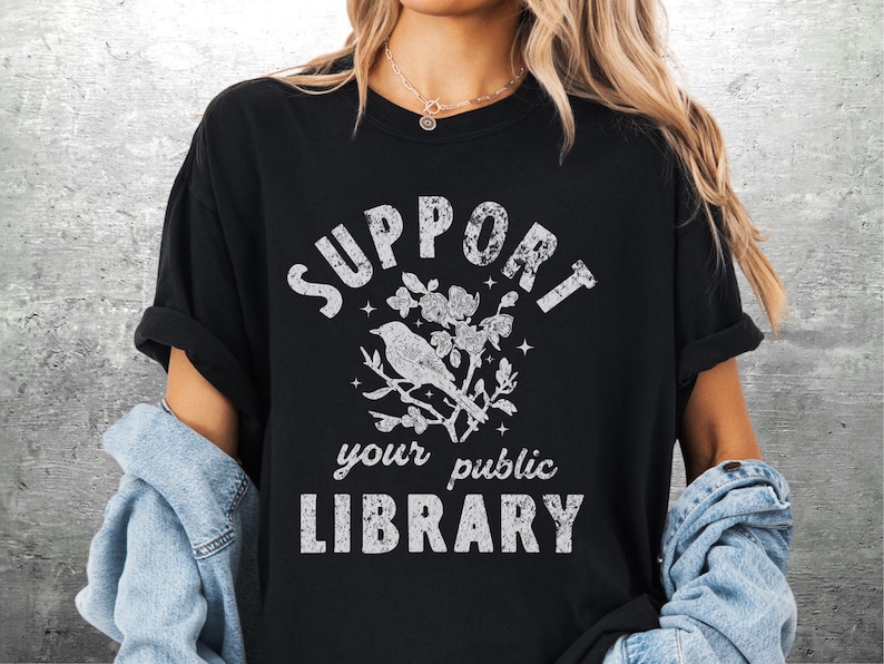 May include: Black t-shirt with the text "SUPPORT your public LIBRARY" in distressed white lettering. A bird perched on a flowering branch is centered above the text. The shirt has rolled-up sleeves.