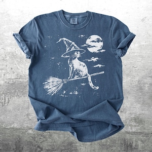 May include: A blue short-sleeve t-shirt featuring a white graphic print of a cat wearing a pointed witch hat, sitting on a flying broomstick against a full moon and clouds. The design has a distressed, weathered texture across the front.