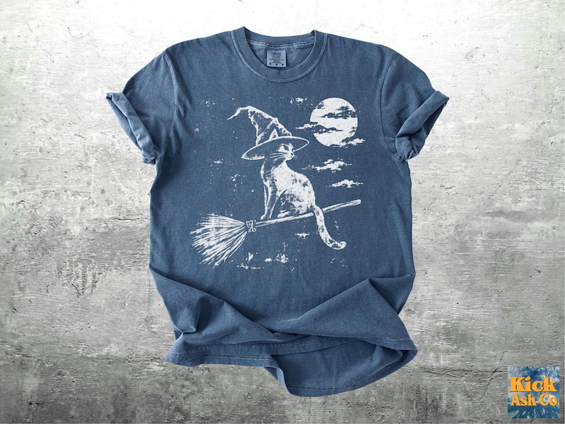 May include: A blue short-sleeve t-shirt featuring a white graphic print of a cat wearing a pointed witch hat, sitting on a flying broomstick against a full moon and clouds. The design has a distressed, weathered texture across the front.