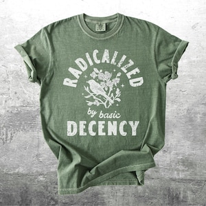 May include: Green t-shirt with rolled sleeves. The shirt features the text "RADICALIZED by basic DECENCY" in white, with a bird and floral graphic. The shirt is displayed on a gray, textured surface.