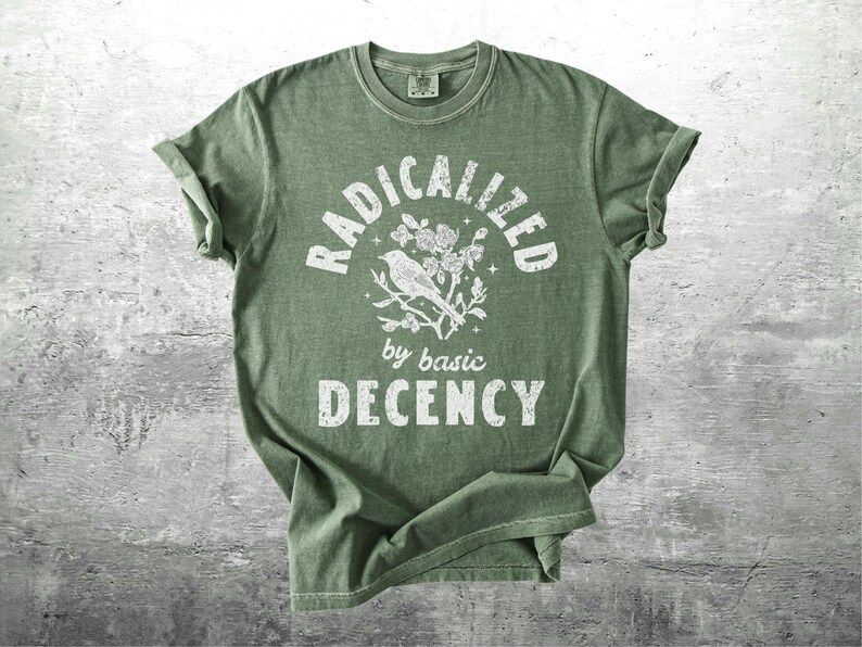 May include: Green t-shirt with rolled sleeves. The shirt features the text "RADICALIZED by basic DECENCY" in white, with a bird and floral graphic. The shirt is displayed on a gray, textured surface.