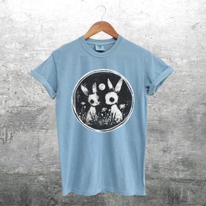 Op de afbeelding: Een lichtblauw t-shirt met een zwart-witte afbeelding van twee konijnen in een cirkel. De konijnen kijken elkaar aan en er is een kleine maan op de achtergrond.