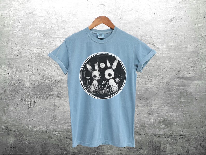 Op de afbeelding: Een lichtblauw t-shirt met een zwart-witte afbeelding van twee konijnen in een cirkel. De konijnen kijken elkaar aan en er is een kleine maan op de achtergrond.