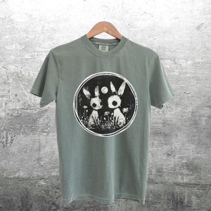 Op de afbeelding: Een saliegroen T-shirt met een zwart-witte afbeelding van twee konijnen in een bosrijke omgeving. De afbeelding bevat een volle maan en een cirkel rond de konijnen.