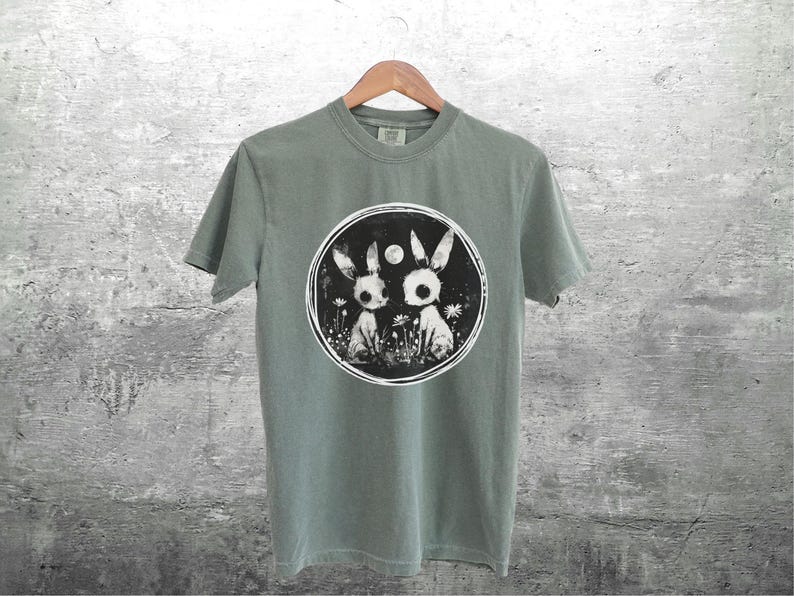 Op de afbeelding: Een saliegroen T-shirt met een zwart-witte afbeelding van twee konijnen in een bosrijke omgeving. De afbeelding bevat een volle maan en een cirkel rond de konijnen.