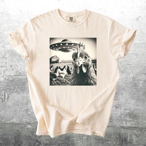 May include: Cream-colored t-shirt featuring a black and white image of a chicken looking at the viewer with a UFO hovering above a farm scene. The shirt has short sleeves and a crew neck.