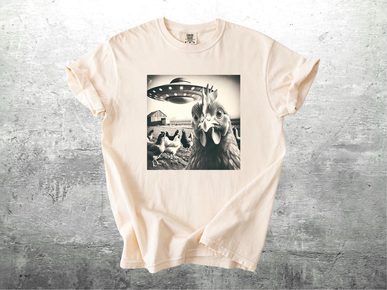 May include: Cream-colored t-shirt featuring a black and white image of a chicken looking at the viewer with a UFO hovering above a farm scene. The shirt has short sleeves and a crew neck.