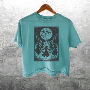 May include: A light teal short-sleeve t-shirt featuring a dark gray rectangular graphic print of four figures in pointed hats dancing in a circle around a bonfire under a large full moon and stars, with the text Kick Ash Co in the bottom corner.