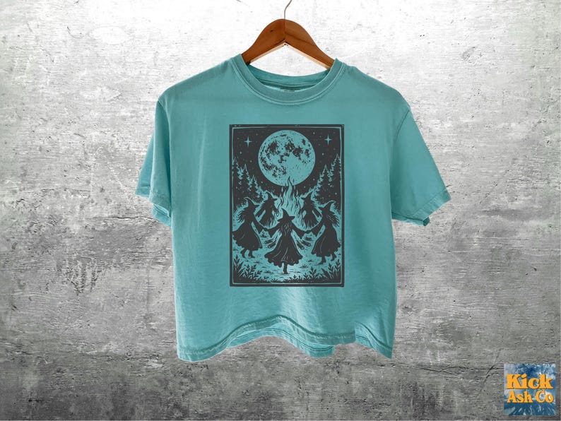 May include: A light teal short-sleeve t-shirt featuring a dark gray rectangular graphic print of four figures in pointed hats dancing in a circle around a bonfire under a large full moon and stars, with the text Kick Ash Co in the bottom corner.