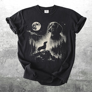 May include: Black t-shirt featuring a dachshund dog design. The graphic includes a full moon, trees, and a silhouette of a dog on a hill. The dog's head is also featured in the design. The shirt is made of a soft material.