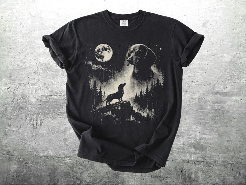 May include: Black t-shirt featuring a dachshund dog design. The graphic includes a full moon, trees, and a silhouette of a dog on a hill. The dog's head is also featured in the design. The shirt is made of a soft material.