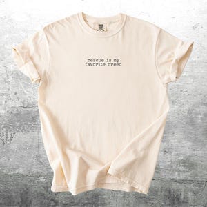 May include: Cream-colored t-shirt with the text "rescue is my favorite breed" in a typewriter font. The shirt is made of soft material and has rolled-up sleeves. The shirt is displayed on a gray, textured surface.