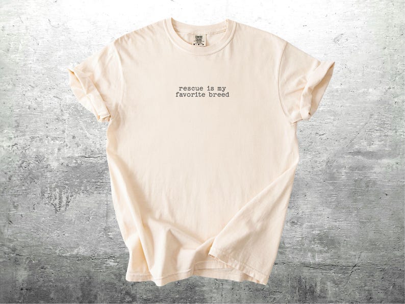 May include: Cream-colored t-shirt with the text "rescue is my favorite breed" in a typewriter font. The shirt is made of soft material and has rolled-up sleeves. The shirt is displayed on a gray, textured surface.