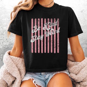 May include: A black short-sleeved t-shirt featuring a graphic design of vertical pink stripes with a distressed texture. Overlaid on the stripes is the white text "Be Kind Stay Weird" in a cursive font. The shirt has a crew neckline and a standard fit.