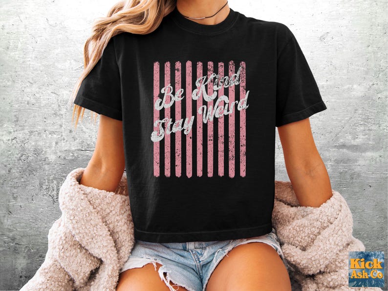 May include: A black short-sleeved t-shirt featuring a graphic design of vertical pink stripes with a distressed texture. Overlaid on the stripes is the white text "Be Kind Stay Weird" in a cursive font. The shirt has a crew neckline and a standard fit.
