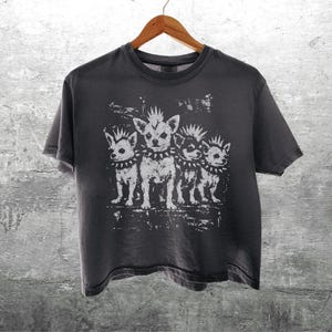May include: Dark gray t-shirt with a distressed graphic of four chihuahua dogs with mohawks and studded collars. The shirt is hanging on a wooden hanger against a textured gray wall.
