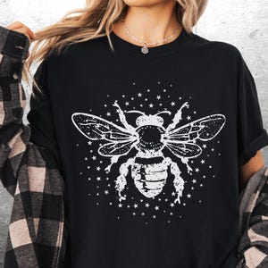 May include: Black crew neck t-shirt featuring a white graphic of a bee with detailed wings and body, surrounded by small stars. The shirt has short sleeves and a relaxed fit, suitable for casual wear.
