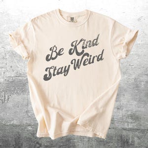 May include: Cream-colored t-shirt with the phrase "Be Kind Stay Weird" printed in a distressed, dark gray font. The shirt is folded, showcasing the rolled sleeves and soft fabric. The background is a textured gray surface.