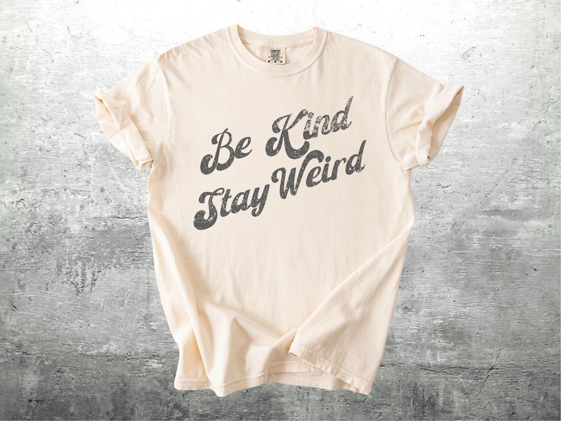 May include: Cream-colored t-shirt with the phrase "Be Kind Stay Weird" printed in a distressed, dark gray font. The shirt is folded, showcasing the rolled sleeves and soft fabric. The background is a textured gray surface.