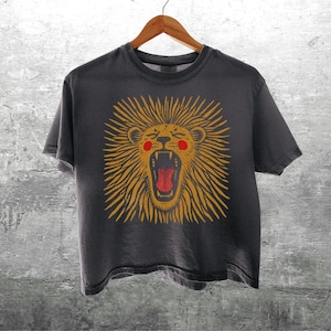 May include: Dark gray t-shirt with a roaring lion graphic. The lion has a yellow mane, red mouth, and pink cheeks. The shirt is hanging on a wooden hanger against a gray concrete wall.