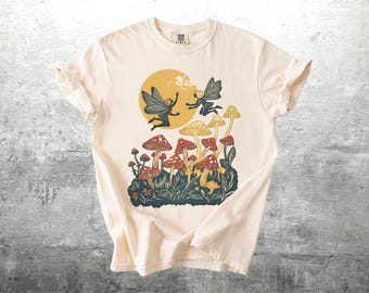 May include: A cream-colored t-shirt with a graphic print of two fairies flying over a field of mushrooms. The mushrooms are red and white with yellow tops. The fairies are wearing brown and white clothing and have brown hair.