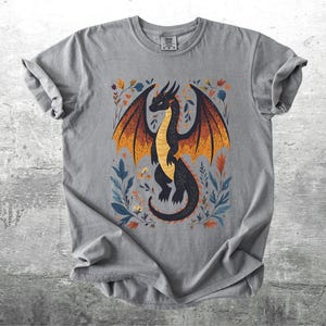 May include: Gray t-shirt with a black dragon illustration with orange and yellow wings. The dragon is surrounded by floral designs in shades of blue, green, and orange.