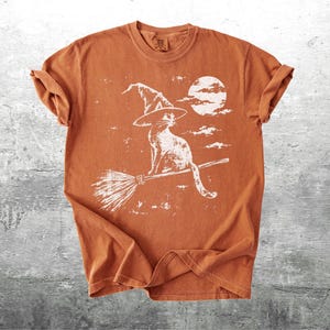 May include: A burnt orange short-sleeve t-shirt featuring a white distressed graphic print of a cat wearing a pointed witch hat, sitting on a broomstick in front of a full moon and scattered clouds. The shirt has a crew neckline and short sleeves.
