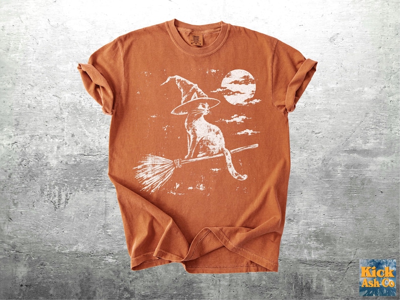 May include: A burnt orange short-sleeve t-shirt featuring a white distressed graphic print of a cat wearing a pointed witch hat, sitting on a broomstick in front of a full moon and scattered clouds. The shirt has a crew neckline and short sleeves.