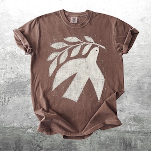 May include: A brown short-sleeve t-shirt featuring a large, distressed white graphic print of a bird holding an olive branch in its beak. The shirt has a crew neckline and a relaxed fit, with the graphic centered on the chest area of the garment.
