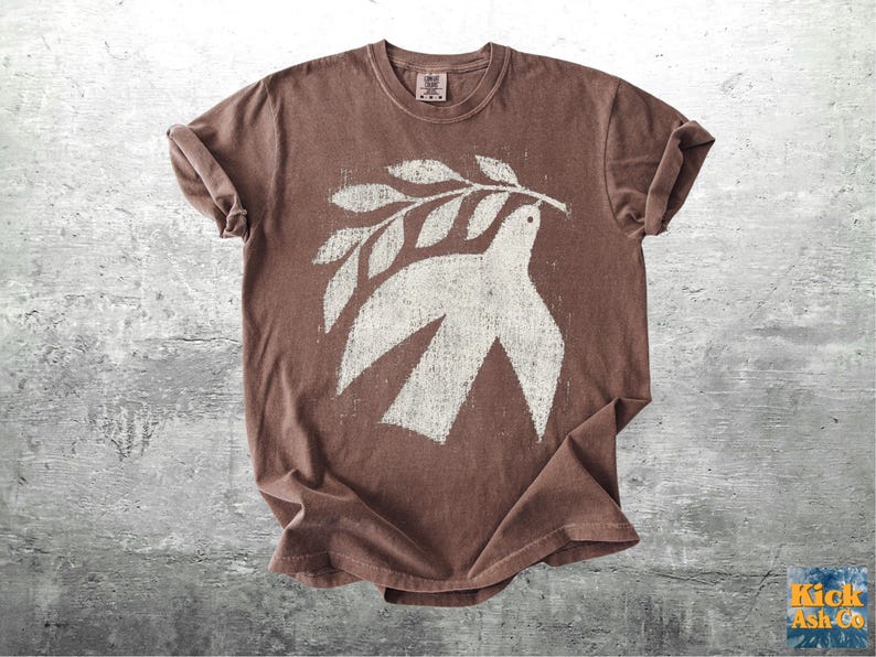 May include: A brown short-sleeve t-shirt featuring a large, distressed white graphic print of a bird holding an olive branch in its beak. The shirt has a crew neckline and a relaxed fit, with the graphic centered on the chest area of the garment.