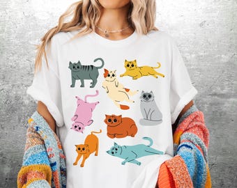 May include: White t-shirt featuring a colorful illustration of various cartoon cats in different poses. The cats are in shades of gray, yellow, orange, pink, and blue. The shirt has a crew neck and short sleeves.