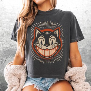 May include: A charcoal gray short-sleeve t-shirt featuring a graphic print of a grinning cat face with wide eyes and a large smile. The cat illustration is outlined in orange and surrounded by radiating lines. The shirt has a crew neckline.