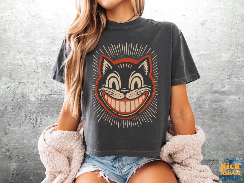 May include: A charcoal gray short-sleeve t-shirt featuring a graphic print of a grinning cat face with wide eyes and a large smile. The cat illustration is outlined in orange and surrounded by radiating lines. The shirt has a crew neckline.