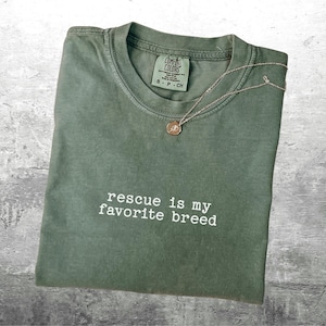 May include: Olive green t-shirt with the phrase "rescue is my favorite breed" in white. A gold-colored pendant necklace rests on the shirt. The shirt has a crew neck and a tag at the neckline.