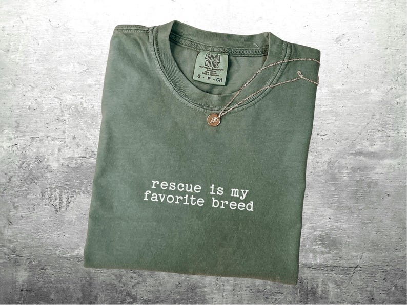 May include: Olive green t-shirt with the phrase "rescue is my favorite breed" in white. A gold-colored pendant necklace rests on the shirt. The shirt has a crew neck and a tag at the neckline.