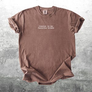 May include: A brown t-shirt with the phrase "rescue is my favorite breed" printed in white. The shirt has a crew neck and short sleeves, with the sleeves rolled up. The fabric has a slightly faded appearance.