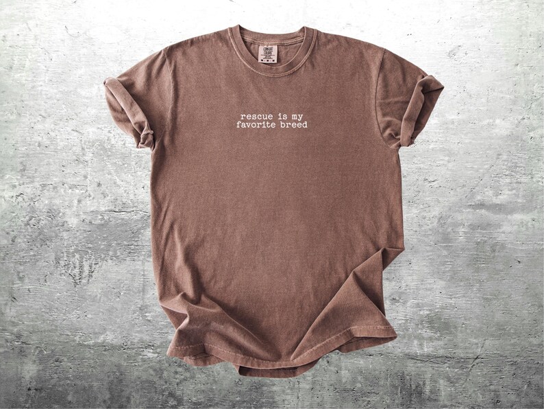 May include: A brown t-shirt with the phrase "rescue is my favorite breed" printed in white. The shirt has a crew neck and short sleeves, with the sleeves rolled up. The fabric has a slightly faded appearance.