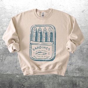 May include: Beige sweatshirt with a blue graphic of a sardine can. The can contains four fish and the text "Sardines Wild Caught in Olive Oil". The sweatshirt has long sleeves and a crew neck.