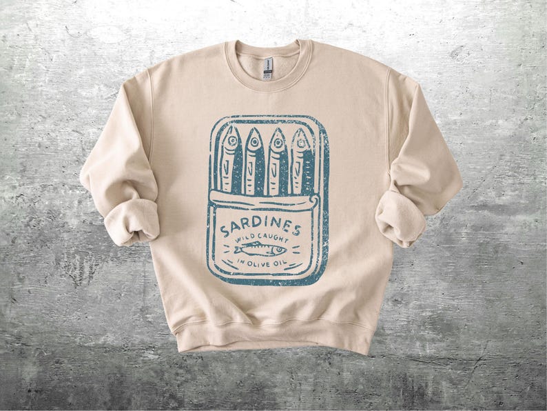 May include: Beige sweatshirt with a blue graphic of a sardine can. The can contains four fish and the text "Sardines Wild Caught in Olive Oil". The sweatshirt has long sleeves and a crew neck.