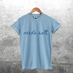 May include: A light blue t-shirt with the text "needs salt" written in a handwritten style.