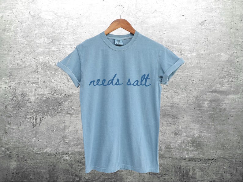 May include: A light blue t-shirt with the text "needs salt" written in a handwritten style.