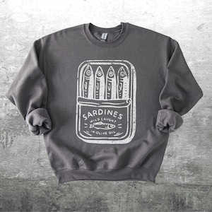 May include: Dark gray sweatshirt featuring a white graphic of a sardine can with the text "Sardines Wild Caught in Olive Oil." The design includes stylized fish illustrations. The sweatshirt has a crew neck and long sleeves.