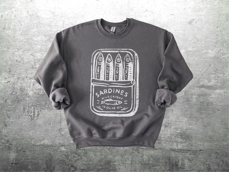 May include: Dark gray sweatshirt featuring a white graphic of a sardine can with the text "Sardines Wild Caught in Olive Oil." The design includes stylized fish illustrations. The sweatshirt has a crew neck and long sleeves.