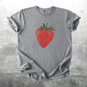 May include: A gray t-shirt featuring a large, red strawberry graphic with green leaves. The strawberry has white dotted details. The shirt is made of a soft material and has rolled-up sleeves.