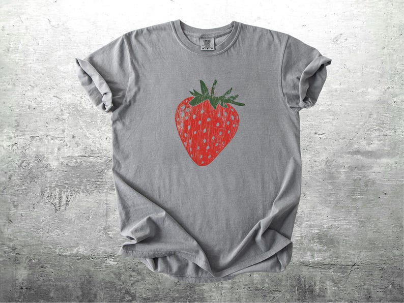 May include: A gray t-shirt featuring a large, red strawberry graphic with green leaves. The strawberry has white dotted details. The shirt is made of a soft material and has rolled-up sleeves.