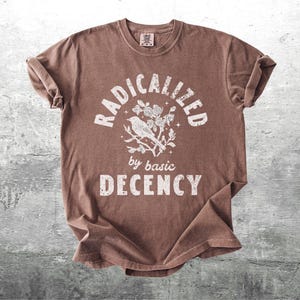 May include: Brown t-shirt with the words "RADICALIZED" above a bird and floral design, with "by basic DECENCY" below. The shirt is made of a soft material and has a crew neck.