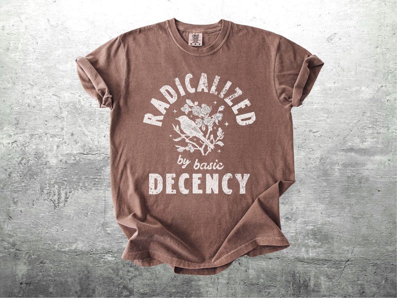 May include: Brown t-shirt with the words "RADICALIZED" above a bird and floral design, with "by basic DECENCY" below. The shirt is made of a soft material and has a crew neck.