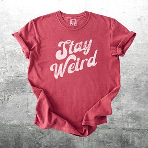 K&ouml;nnte beinhalten: Ein rotes T-Shirt mit dem wei&szlig;en Schriftzug "Stay Weird" in einer stilisierten Schriftart. Die &Auml;rmel sind hochgekrempelt. Das T-Shirt liegt auf einer grauen, strukturierten Oberfl&auml;che. Der Stil ist l&auml;ssig und vintage.