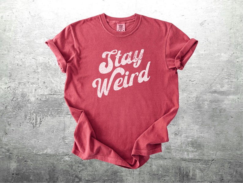 K&ouml;nnte beinhalten: Ein rotes T-Shirt mit dem wei&szlig;en Schriftzug "Stay Weird" in einer stilisierten Schriftart. Die &Auml;rmel sind hochgekrempelt. Das T-Shirt liegt auf einer grauen, strukturierten Oberfl&auml;che. Der Stil ist l&auml;ssig und vintage.