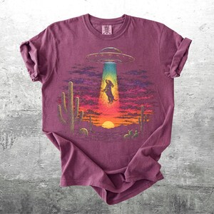 May include: A maroon short-sleeve t-shirt featuring a graphic of a dachshund being abducted by a UFO in a desert landscape at sunset. The design includes a bright beam of light, saguaro cacti, and a vibrant sky with purple and orange hues.